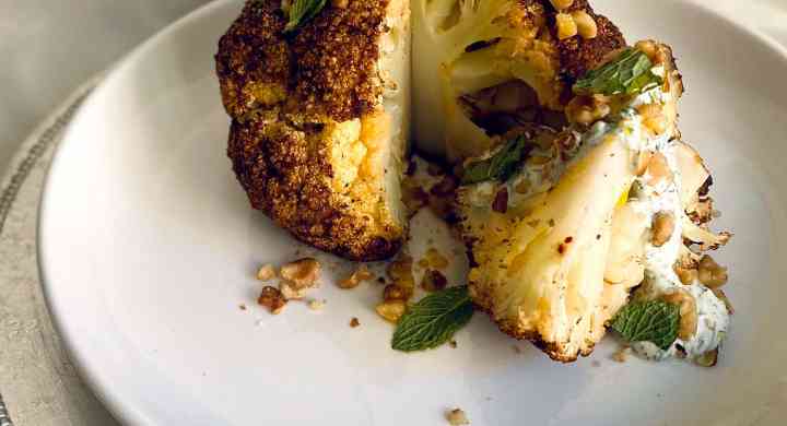 Whole roasted cauliflower with wedge cut out topped with herbs and yogurt sauce on a white plate