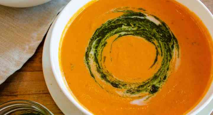 Two white bowls of roasted tomato soup sit on a wooden top table, with fresh whole tomatoes and a jar of pesto beside.